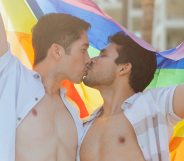 Two gay people kissing in open shirts while holding an LGBTQ+ Pride flag above their heads