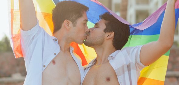 Two gay people kissing in open shirts while holding an LGBTQ+ Pride flag above their heads