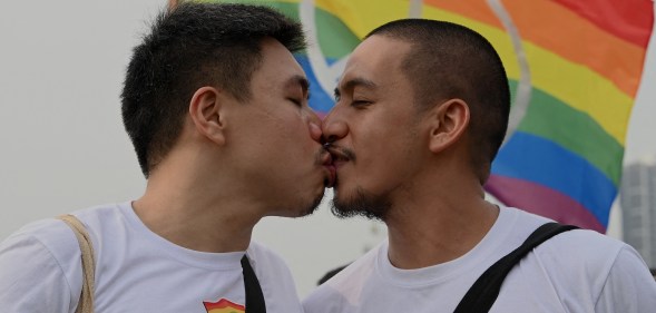 A queer or gay couple sharing a kiss in front of an LGBTQ Pride flag