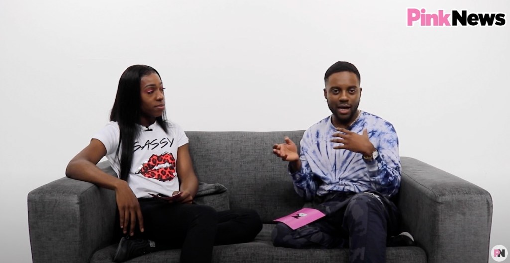 This is an image of two Black men. On the left, he is wearing a white tshirt and has long black hair. On the right, he is wearing a tyedye shirt. They are both sitting on a grey couch in a white room.
