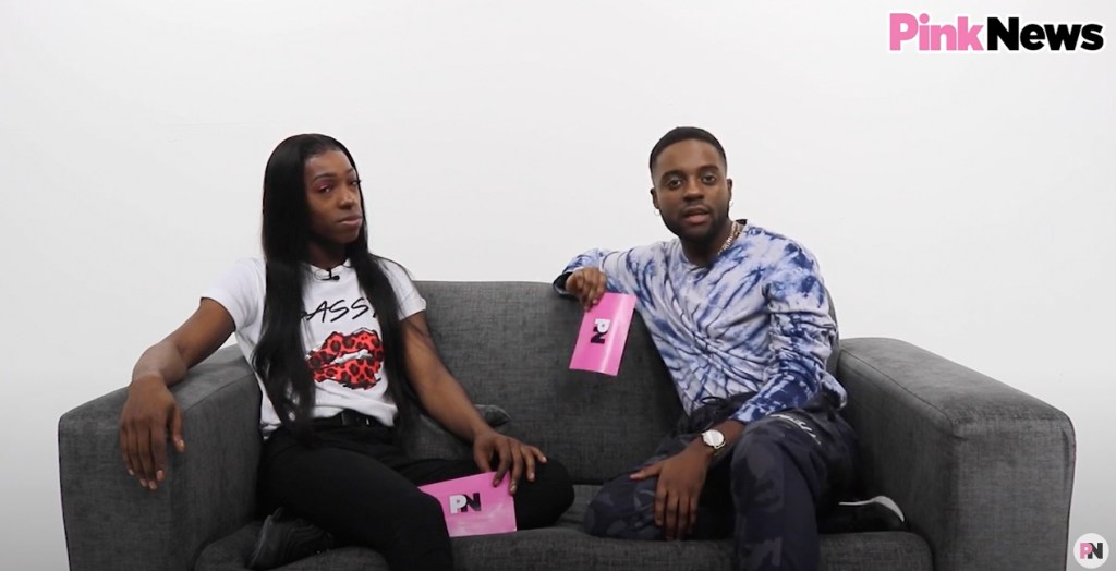 This is an image of two Black men sitting on a couch in a white room. The man on the left has long black hair and is wearing a white shirt. The man on the right has short hair and is wearing a tie dye shirt.