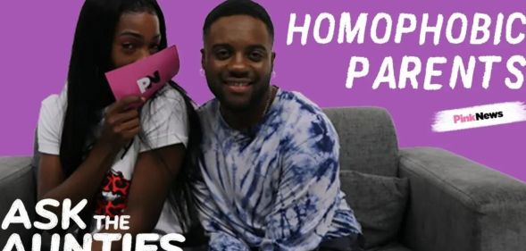This is an image of two Black men sitting on a couch. The person on the left has long black hair and is holding a pink card in front of their face. The other man is sitting next to them and is wearing a tie-dyed shirt. There is white letters spelling Ask the Aunties on the lower left of the screen and Homophobic Parents in the upper right hand of the screen.