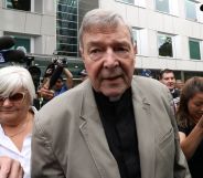 Photo of Cardinal George Pell wearing a gret suit jacket over a black shirt as he leaves Victoria county court in Melbourne surrounded by photographers
