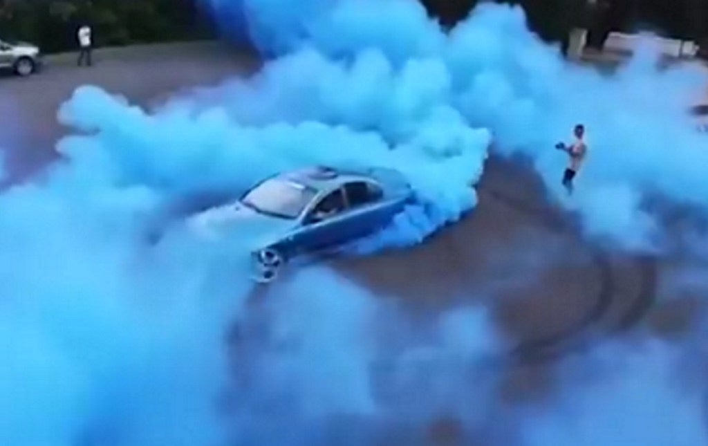 A man drives a car to perform a gender reveal while another man films it.