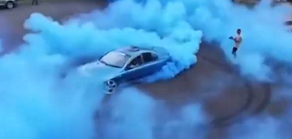 A man drives a car to perform a gender reveal while another man films it.