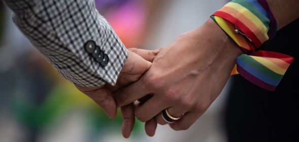 A close-up of a same-sex couple holding hands, one has a rainbow ribbon wrapped around their wrist