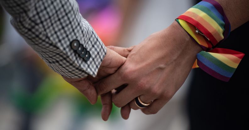 A close-up of a same-sex couple holding hands, one has a rainbow ribbon wrapped around their wrist
