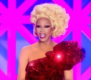 RuPaul in a one-shouldered sequinned red dress and a blonde updo, smiling behind the Drag Race UK judging table