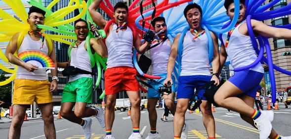 A group of men in white vests and shorts. Each holds balloons in a different colour of the rainbow.