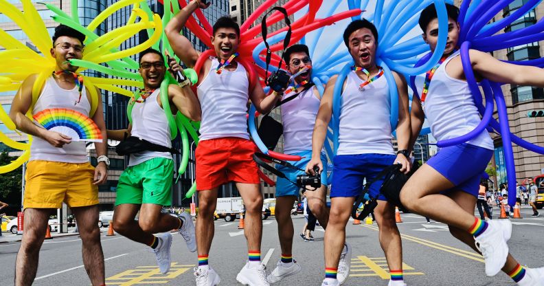 A group of men in white vests and shorts. Each holds balloons in a different colour of the rainbow.