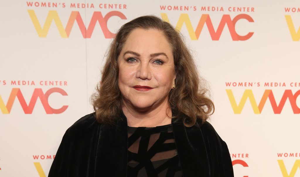 A photo shows actor Kathleen Turner wearing a black dress and smiling at the camera during a press event at the Women's Media Awards