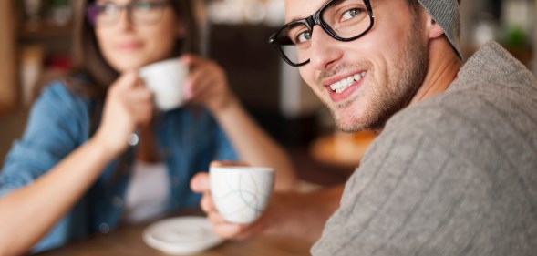 A man smiles at the camera while his girlfriend drinks coffee