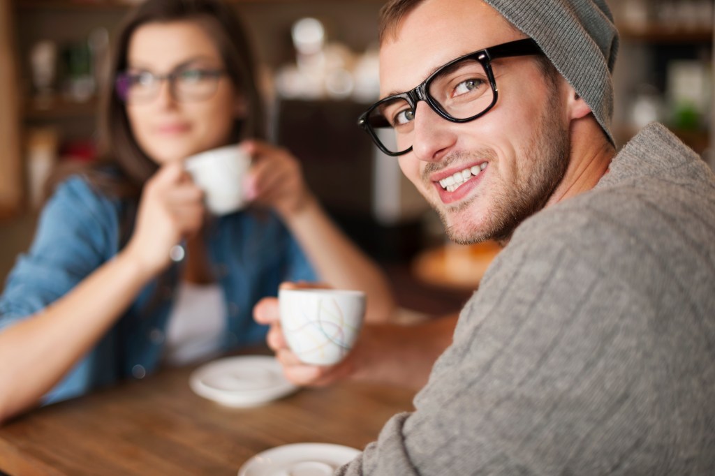 A man smiles at the camera while his girlfriend drinks coffee