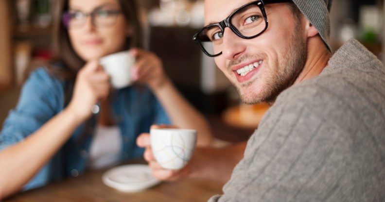 A man smiles at the camera while his girlfriend drinks coffee
