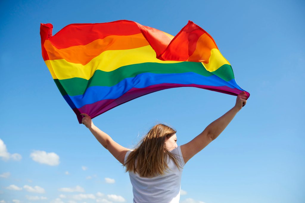 Woman waving an LGBT+ Pride flag against a blue sky
