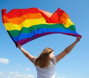 Woman waving an LGBT+ Pride flag against a blue sky