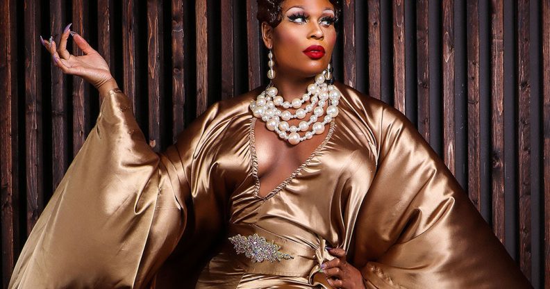 A promo of RuPaul's Drag Race showing drag queen Peppermint wearing a gold dress and over-the-top white pearl necklace posing in front of a dark wooden background