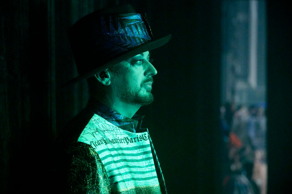 The singer Boy George stands side-on wearing a hat with a green light shining on him