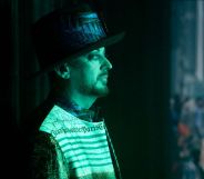 The singer Boy George stands side-on wearing a hat with a green light shining on him