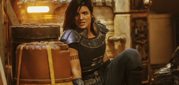 Gina Carano as Cara Dune in The Mandalorian in a body-armour style outfit, hiding behind a barrell