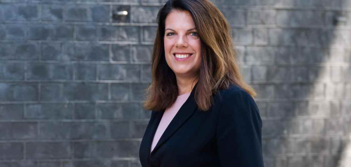 Photo of Conservative MP Caroline Nokes wearing a black suit standing in front of a grey brick wall