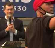 Pete Buttigieg speaking into a microphone as a man wearing a red MAGA cap stands in front of him speaking into his phone, livestreaming