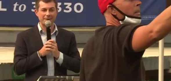 Pete Buttigieg speaking into a microphone as a man wearing a red MAGA cap stands in front of him speaking into his phone, livestreaming
