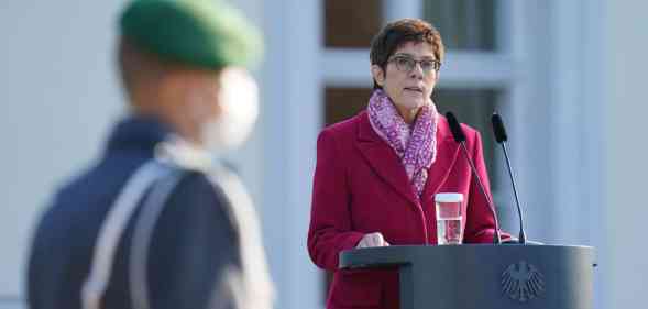 Annegret Kramp-Karrenbauer stands at a podium in a coat. A blurred serviceman is seen in the foreground.