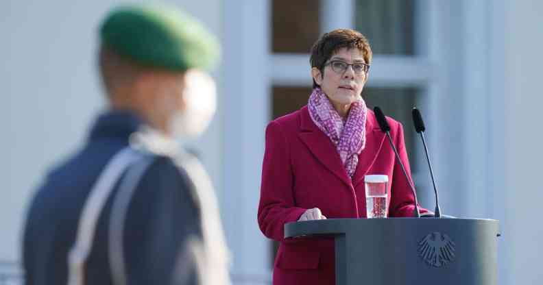 Annegret Kramp-Karrenbauer stands at a podium in a coat. A blurred serviceman is seen in the foreground.
