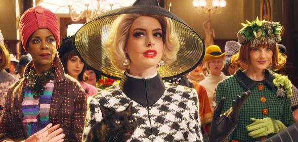 Anne Hathaway in The Witches, wearing a big black hat, blonde hair, and carrying a black cat in a grand ballroom