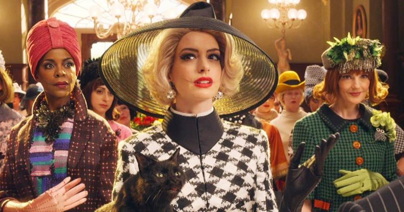 Anne Hathaway in The Witches, wearing a big black hat, blonde hair, and carrying a black cat in a grand ballroom