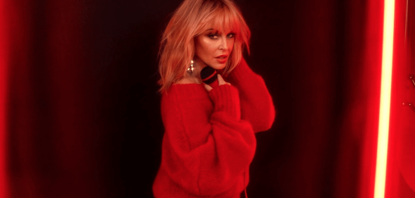 Kylie Minogue looking over her shoulder in a red jumper, a soft red neon lighting her face. She's holding a microphone