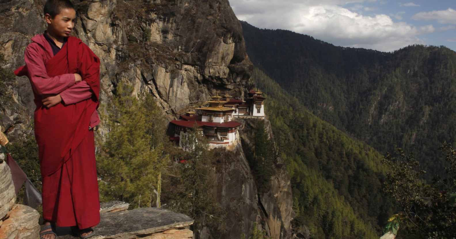 A monk stands on the mountaintops, a monastery in view behind him on the cliffs