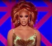 RuPaul in a gold dress with copper hair sitting in front of the Drag Race UK judges' panel backdrop