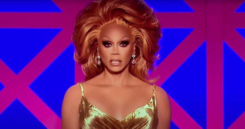 RuPaul in a gold dress with copper hair sitting in front of the Drag Race UK judges' panel backdrop