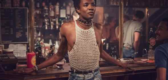 Omari Douglas as Roscoe, leaning against a pub bar, wearing acid-washed jeans and a string vest