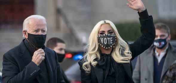 Joe Biden and Lady Gaga, both wearing masks, wave while wearing dark coats