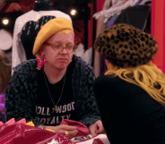 Ginny Lemon and Bimini Bon Boulash holding hands sitting across a pink werk room table