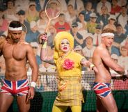 Ginny Lemon in a yellow suit holding a tennis racket, flanked by two Brit crew wearing just Union Jack boxers