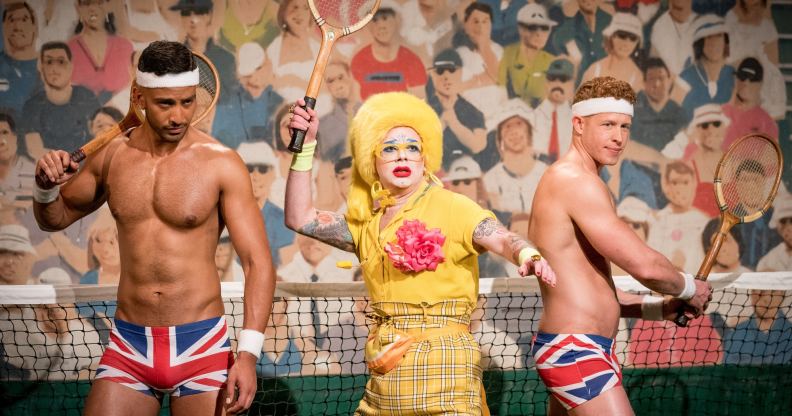 Ginny Lemon in a yellow suit holding a tennis racket, flanked by two Brit crew wearing just Union Jack boxers