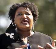 Close-up of Stacey Abrams speaking. A black face mask with 'VOTE' in bold white text is hanging from one ear.