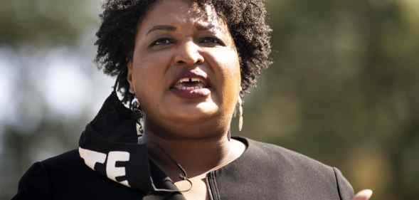 Close-up of Stacey Abrams speaking. A black face mask with 'VOTE' in bold white text is hanging from one ear.
