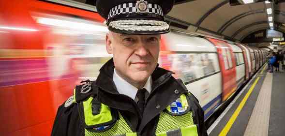 Adrian Hanstock, a police officer wearing a yellow h-vis vest, black jacket and tie and a police cap, standing on a Tube platform as a train speeds past