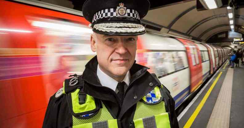 Adrian Hanstock, a police officer wearing a yellow h-vis vest, black jacket and tie and a police cap, standing on a Tube platform as a train speeds past