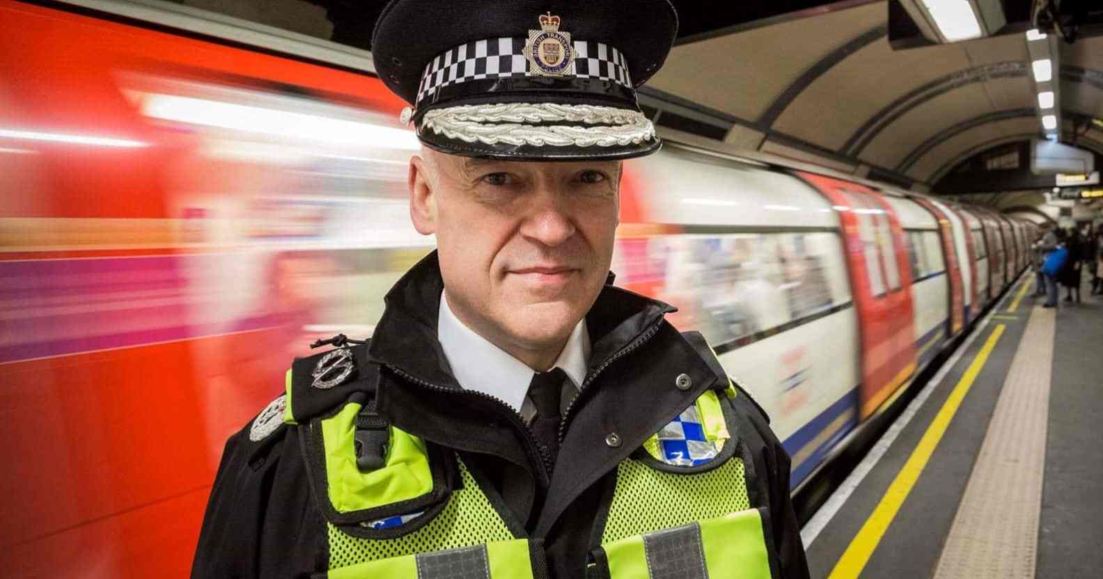 Adrian Hanstock, a police officer wearing a yellow h-vis vest, black jacket and tie and a police cap, standing on a Tube platform as a train speeds past