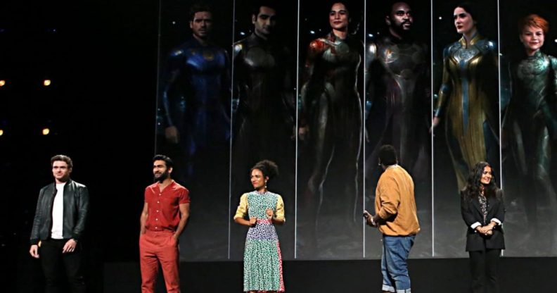 Richard Madden, Kumail Nanjiani, Lauren Ridloff, Brian Tyree Henry, and Salma Hayek on a stage infant of images of them in superhero costume