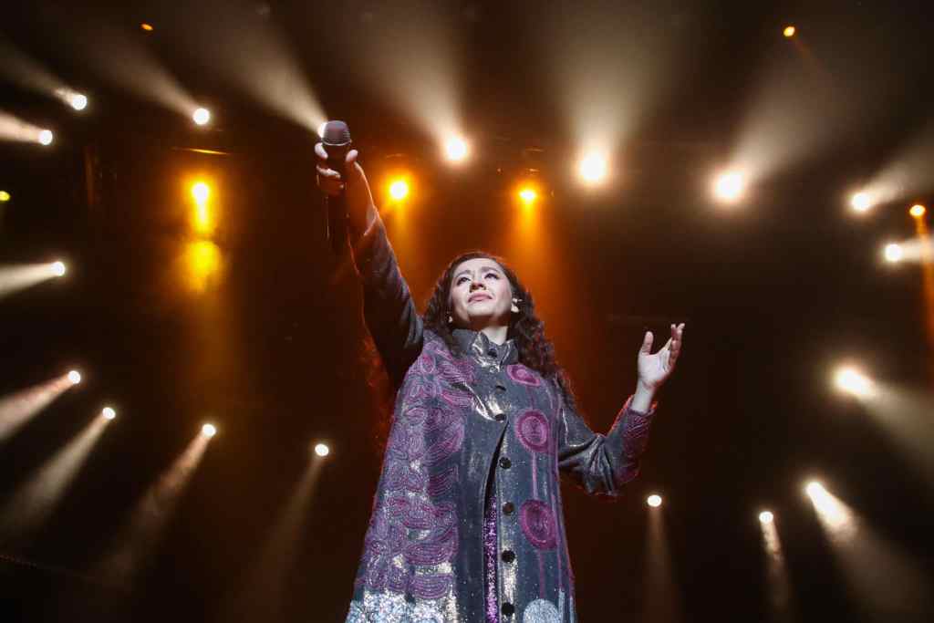Manizha Sangin holds her microphone in the air against a constellation of spotlights