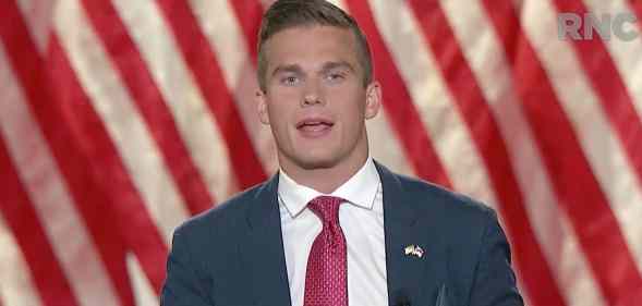 Madison Cawthorn addresses the virtual 2020 Republican National Convention, wearing a navy suit, white shirt and red tie against the stripes of the American flag