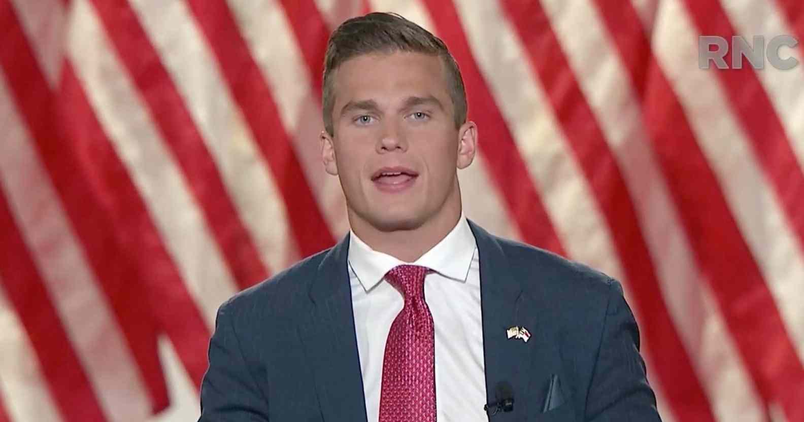 Madison Cawthorn addresses the virtual 2020 Republican National Convention, wearing a navy suit, white shirt and red tie against the stripes of the American flag