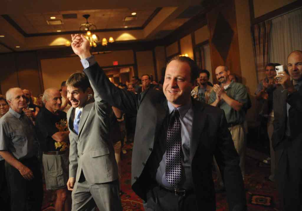 Jared Polis (R) and Marlon Reis hold hands as people clap behind them